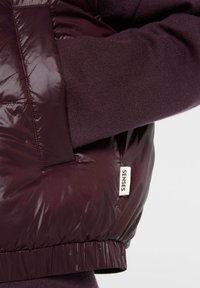 Burgunderfarbene Pufferjacke mit glänzender Textur, gerippten Bündchen und einem "SENSES"-Label an der Seite. Weicher Strickstoff unter den Ärmeln.