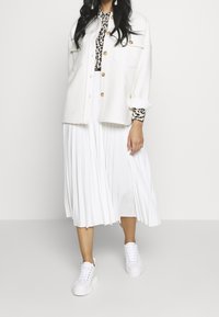 Mujer vestida con una chaqueta blanca con botones, una camiseta de estampado de leopardo, una falda midi blanca plisada y zapatillas deportivas blancas con plataforma, de pie sobre un fondo liso.