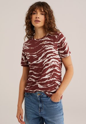 Donna con capelli ricci che indossa una maglietta a maniche corte a righe bordeaux e bianche e jeans blu, una mano in tasca, sfondo neutro.