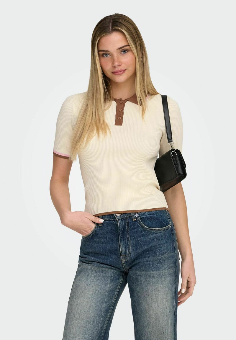 Jeune femme aux longs cheveux blonds portant un polo crème avec des bordures marron, un jean bleu et un sac à bandoulière noir, posant avec une main dans la poche.