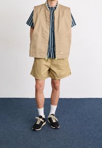 Osoba v béžové vestě, odpovídajících šortkách, modré pruhované košili s krátkým rukávem, bílých ponožkách a černých teniskách stojí na modrém koberci.