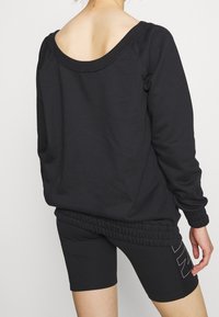 Svart sweatshirt med vid halsringning och ribbad nederkant, kombinerad med svarta cykelshorts som har en vit logotypkontur på sidan.