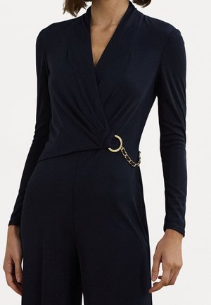 Kvinde iført en sort langærmet slå-om-jumpsuit med en guldring og kædedetalje ved taljen, stående mod en hvid baggrund.