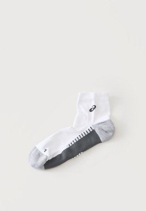 Weißer Knöchelsocken mit grauer Ferse und Spitze, schwarzem Sohlenmuster und kleinem schwarzem Logo nahe dem Bündchen auf einem schlichten hellen Hintergrund.