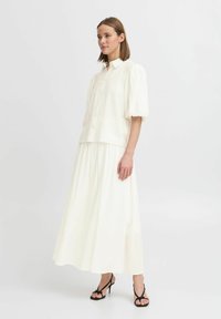 Blouse blanche à manches bouffantes avec un devant à boutons, associée à une longue jupe maxi blanche fluide. Complétée par des sandales noires à lanières. Texture lisse.