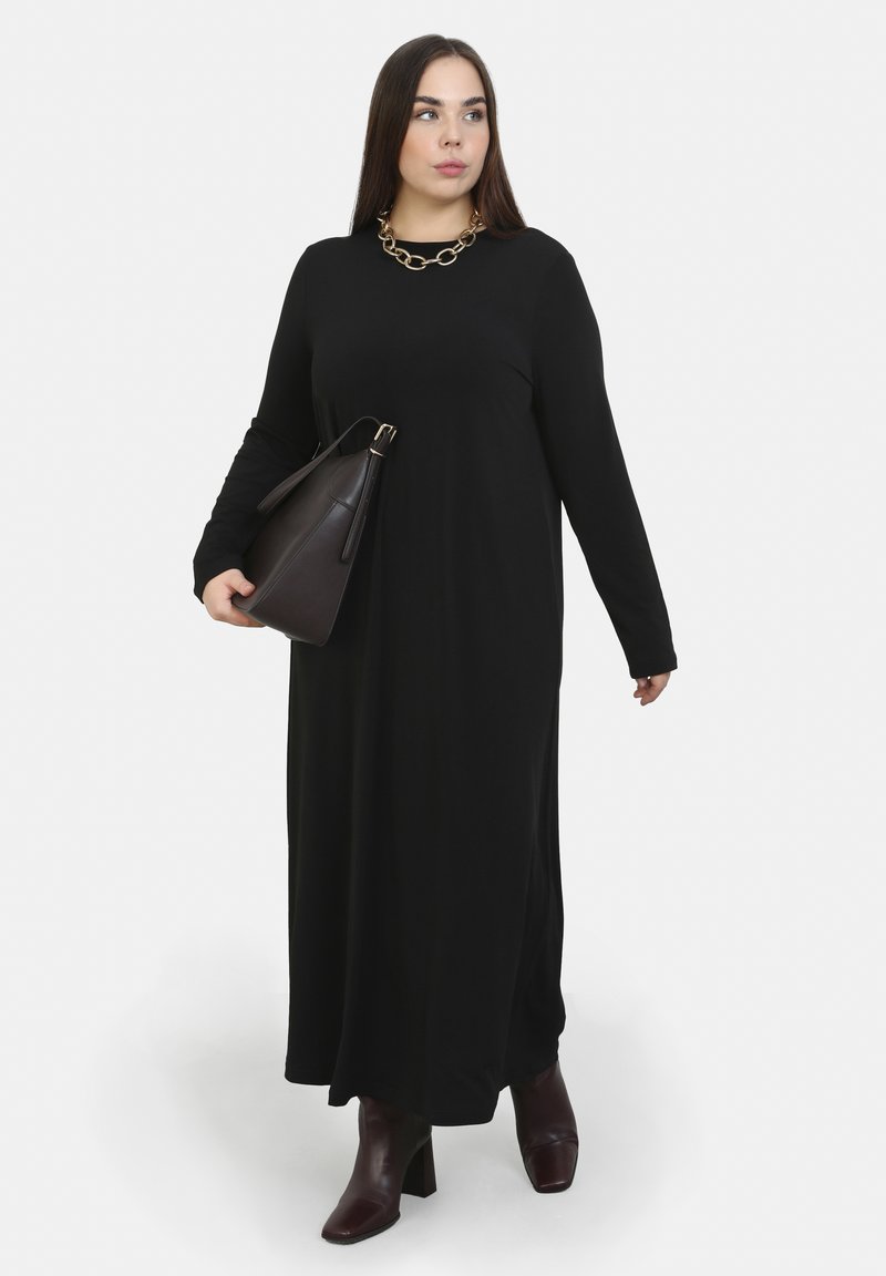 Schwarzes Langarmkleid mit rundem Ausschnitt, kombiniert mit einer dunklen Handtasche. Das Model trägt kastanienbraune Absatzstiefel und eine dicke goldene Halskette.