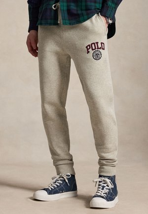 Mann trägt beige Sweatpants mit Polo-Logo am Oberschenkel, blaue High-Top-Sneaker und ein grün-navy kariertes Hemd mit der Hand in der Tasche.