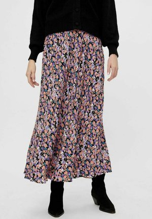 Vrouw die een zwarte cardigan draagt en een lange bloemenrok met roze, paarse en blauwe bloemen, gecombineerd met zwarte laarzen met hak.