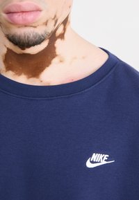 Námořnická modrá bavlněná mikina s ribárovým výstřihem do krku a bílým vyšitým logem Nike na hrudi. Hladká textura, minimalistický design.