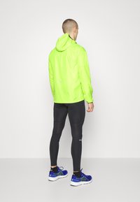 Veste légère vert fluo avec capuche, associée à un legging noir ajusté et des chaussures de sport bleues. Accents réfléchissants sur la veste.