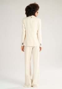 Abito color crema con righe sottili, composto da un blazer sartoriale con un bottone solo al colletto e pantaloni a gamba larga per un aspetto formale.