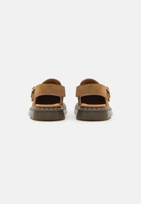 Sandales en suède marron avec un talon ouvert, une sangle réglable et une semelle en caoutchouc noire. Des coutures jaunes soulignent la semelle.