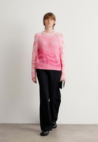 Model loopt in een roze geblokte trui, wijde zwarte broek, zwarte schoenen en houdt een kleine zwart-wit clutch vast.