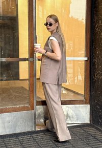 Beige sleeveless vest over a white shirt, paired with wide-legged beige pants. Holds a coffee cup, stands next to a glass door.
