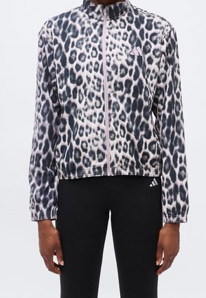 Mujer con una chaqueta con cremallera con estampado de leopardo blanco y negro y mallas negras con un pequeño logo blanco en el muslo izquierdo.
