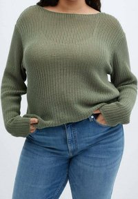 Groene, chunky gebreide trui met een wijde halslijn en lange mouwen; gedragen met strakke blauwe jeans, die een relaxte pasvorm en gestructureerd patroon tonen.