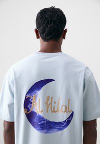 Gaiši zils kokvilnas T-krekls ar lielu zilu pusmēness grafiku, kurā ir mākoņu detaļas un zelta teksts "Al Hilal". Attēlotā aizmugures skats.