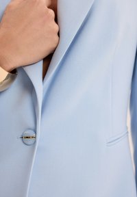 Jasnoniebieska marynarka z teksturowanej tkaniny, z eleganckim klapem, okrągłym guzikiem ze złotym akcentem oraz detalem bocznej kieszeni.