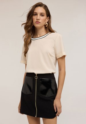 Top beige a maniche corte con bordi neri, abbinato a una mini gonna nera con tasche frontali lucide e una zip centrale.