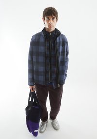 Marineblauwe en grijze ruitjes zip-up jack, gelaagd over een ruitjeshemd, gecombineerd met bordeauxrode corduroy broek en een paarse en grijze tote bag. Zilveren sneakers.