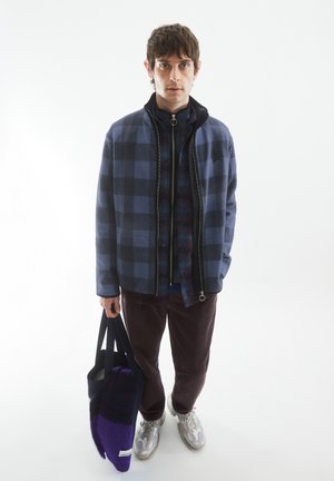 Marineblauwe en grijze ruitjes zip-up jack, gelaagd over een ruitjeshemd, gecombineerd met bordeauxrode corduroy broek en een paarse en grijze tote bag. Zilveren sneakers.
