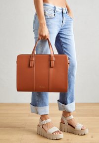 Sac en toile marron en cuir avec des poignées doubles, de forme rectangulaire et des accents cousus. Associé à un jean bleu clair à ourlets et des sandales à plateforme beiges.