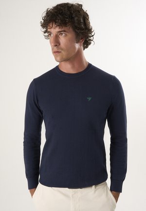Dunkelblauer Langarmpullover in Stricktextur mit Rundhalsausschnitt. Verfügt über ein kleines grünes Logo auf der Brust. Kombinierbar mit hellen Hosen.