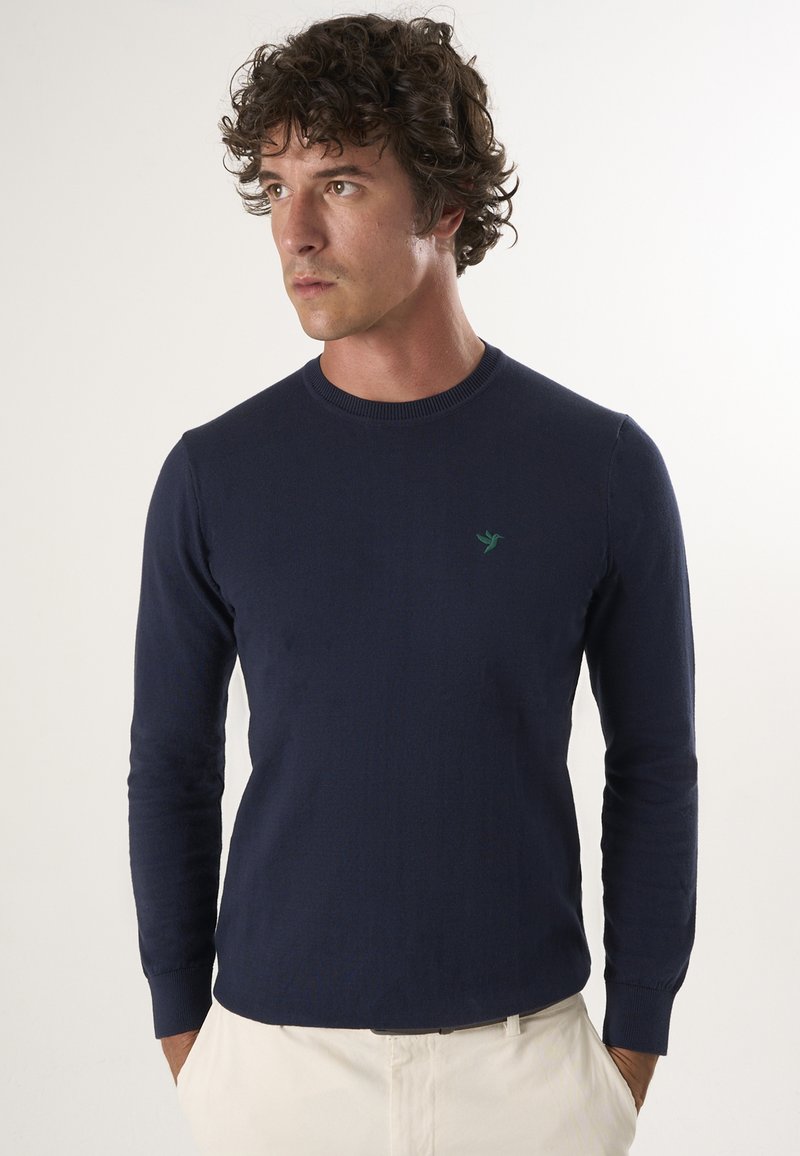 Marineblauwe lange mouwen trui in een gebreide structuur met een ronde hals. Heeft een klein groen logo op de borst. Combineer met lichte broeken.