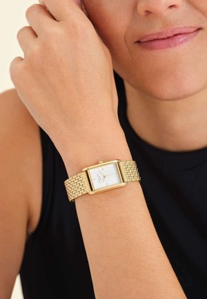 Montre rectangulaire en or avec un cadran blanc et des marqueurs d'heure simples, dotée d'un bracelet en maille texturée, portée sur un poignet à peau claire.