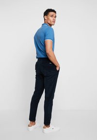 Sininen polo-paita valkoisilla yksityiskohdilla, tummat housut ja valkoiset tennarit. Malli poseeraa yksi käsi taskussa, esitellen vaatteen istuvuutta ja tyyliä.