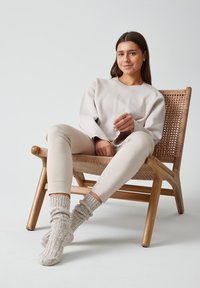 Sudadera gris claro y pantalones ajustados beige, usando calcetines de punto gris claro texturizados, sentado en una silla de mimbre de madera frente a un fondo simple.