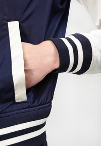 Námořnická bombardovací bunda s bílými kontrastními pruhy. Hladká látka s žebrovanými manžetami. Obsahuje boční kapsu umístěnou na levé straně.