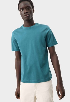 Giovane uomo che indossa una t-shirt a maniche corte color verde acqua e pantaloni bianco sporco, in piedi davanti a uno sfondo chiaro e semplice.