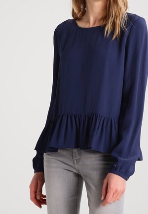 Femme portant un chemisier bleu marine à manches longues avec un col rond et un ourlet à volants, associé à un jean gris clair.