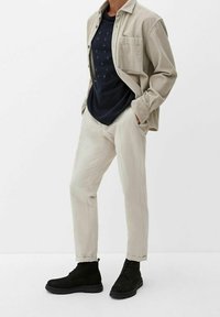 Beige Cord-Hemd über einem dunkelblauen T-Shirt mit Text. Cremefarbene Hose und schwarze Wildleder-Ankle-Boots mit dicker Sohle.