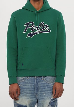 Mann trägt grünen Polo Ralph Lauren Hoodie mit großem Logo auf der Brust und blaue, abgenutzte Jeans, steht vor neutralem Hintergrund.