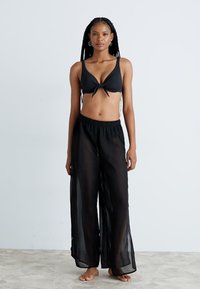 Crni bikini gornji dio s vezanjem sprijeda, uparen s prozirnim crnim širokim hlačama. Glatka tekstura; opušteni kroj za udobnost i prozračnost.