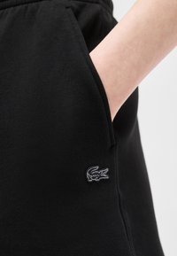 Χέρι στην τσέπη μαύρου παντελονιού με κεντημένο λευκό περίγραμμα λογότυπου κροκόδειλου στο μηρό.