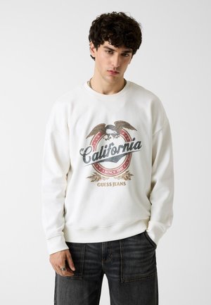 Jeune homme portant un sweat-shirt blanc avec un aigle vintage et le logo "California Guess Jeans", assorti à un jean denim foncé.