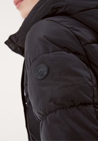 Svart puffjacka med kviltad textur, utrustad med en rund svart logotyp-patch på vänster ärm. Huvudets design med dragkedja framtill.
