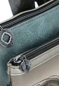 Bolso multicolor con un exterior texturizado en color teal, acentos plateados y cremalleras oscuras. Presenta herrajes metálicos distintivos y detalles en relieve.