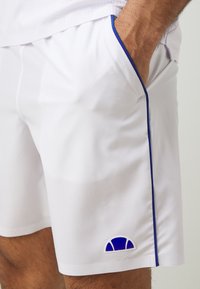 Weiße Shorts aus leichtem Stoff, mit blauen Paspeln und einem kleinen blauen Logodetail an der unteren Seite. Zwei seitliche Taschen sind sichtbar.