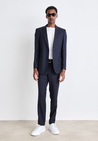 Completo da uomo navy con due bottoni, slim fit, abbinato a una maglietta bianca e sneakers bianche. Gli accessori includono occhiali da sole neri e una cintura nera.