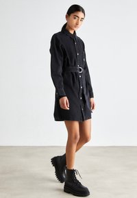 Femme portant une robe-shirt en denim noire ceinturée et des bottes à lacets noires imposantes, se tenant contre un fond clair et uni.