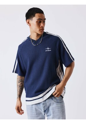 Jeune homme regardant vers le bas portant une chemise bleue texturée à manches courtes avec des rayures blanches et un jean en denim clair, montrant un bras tatoué.