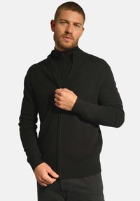 Czarny sweter na zamek z wysokim kołnierzem, ściągaczami na mankietach i dolnej krawędzi. Wykonany z miękkiego, teksturowanego materiału. Noszony częściowo rozpięty.
