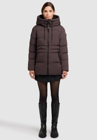 Puffere jakke i mørkebrun med hætte, frontlynlås og to sidelommer. Kviltet design tilføjer tekstur. Bæret over en sort nederdel og strømpebukser.
