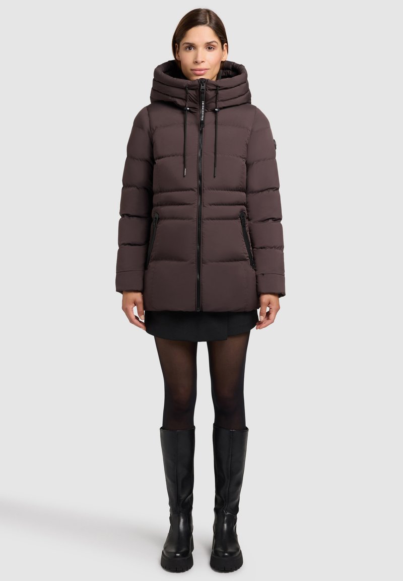 Puffere jakke i mørkebrun med hætte, frontlynlås og to sidelommer. Kviltet design tilføjer tekstur. Bæret over en sort nederdel og strømpebukser.