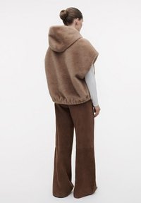 Ruskea fleece-poncho hupulla, pehmeällä pinnalla ja leveällä leikkauksella. Yhdistetty ruskeisiin mokkanahkaisiin leveälahkeisiin housuihin.