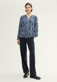 Kwiatowa bluzka z długim rękawem w granatowych i jasnoniebieskich odcieniach, z dekoltem w kształcie litery V i elastycznymi mankietami, zestawiona z ciemnymi spodniami o klasycznym kroju i czarnymi butami.